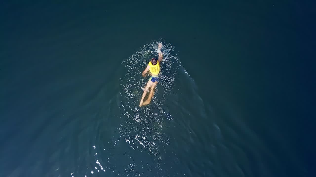 Aerial shot of a boy swimming in clear sea water. Top view of a boy swimming away camera in the middle of the ocean