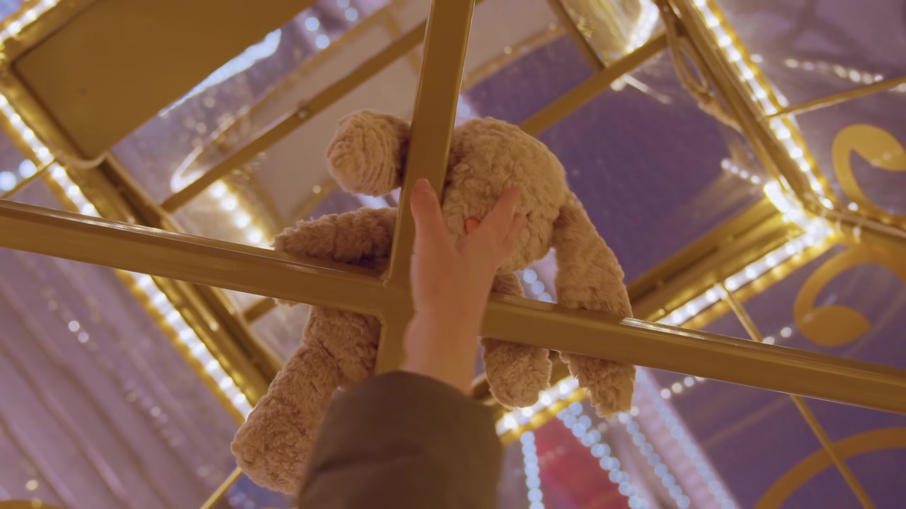 Hand reaching teddy bear inside carousel cage, fair visitor stretching upward under warm marquee lights to retrieve plush prize, closeup of metal lattice, soft fur, hopeful mood nostalgic atmosphere