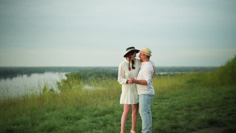 a joyful moment as a husband and wife dance together on a grassy hill beside a tranquil lake. the man wears a white shirt, hat, and jeans, while the woman is dressed in a white dress and black hat