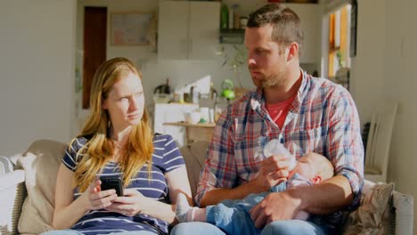 Vista-Frontal-Padre-Caucásico-Adulto-Medio-Alimentando-Leche-A-Su-Bebé-Y-A-Su-Madre-Usando-El-Teléfono-Móvil-En-Casa-4k