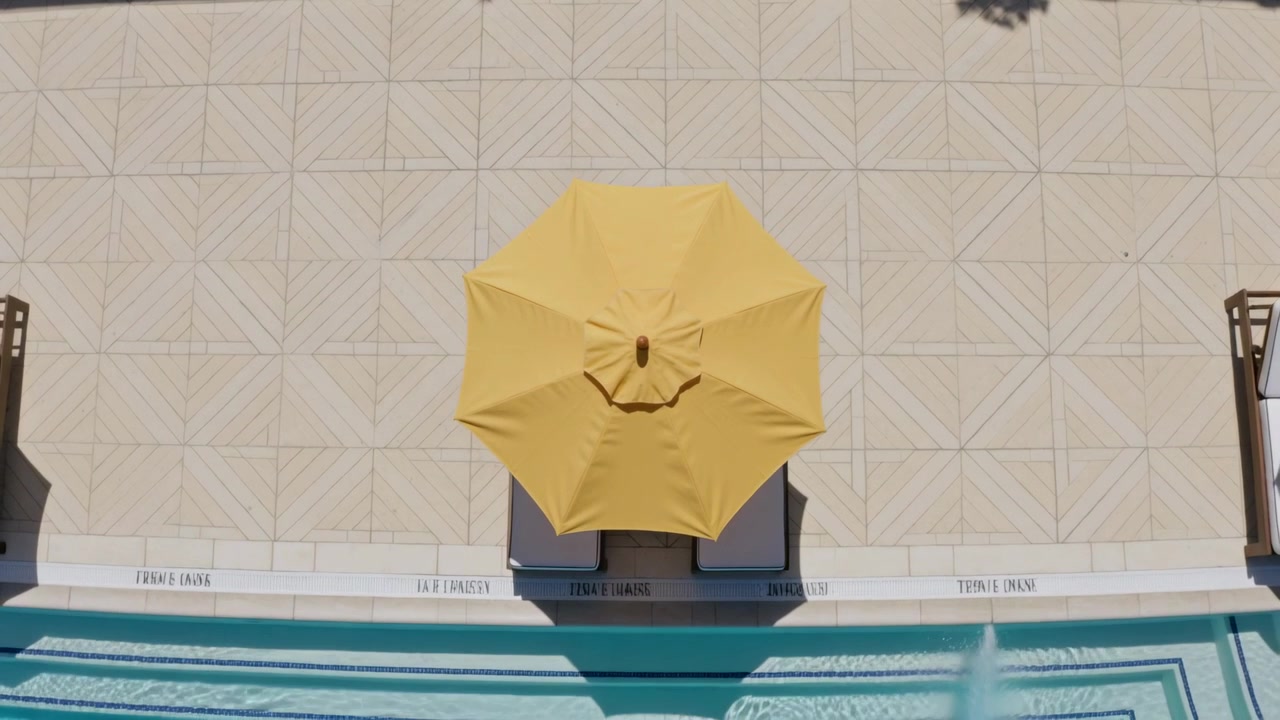 Telecamera aerea che si muove e zooma a bordo piscina per rivelare un ombrellone giallo sopra due lettini e un getto d'acqua che sale.