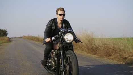 Portrait-of-a-stylish-cool-young-man-in-sunglasses-and-leather-jacket-riding-motorcycle-on-a-road-on-a-sunny-day