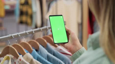 clothing store: female using smartphone with chroma key green screen display. clothes hanger with stylish branded items for retail sale in the background. close up footage of mobile device.