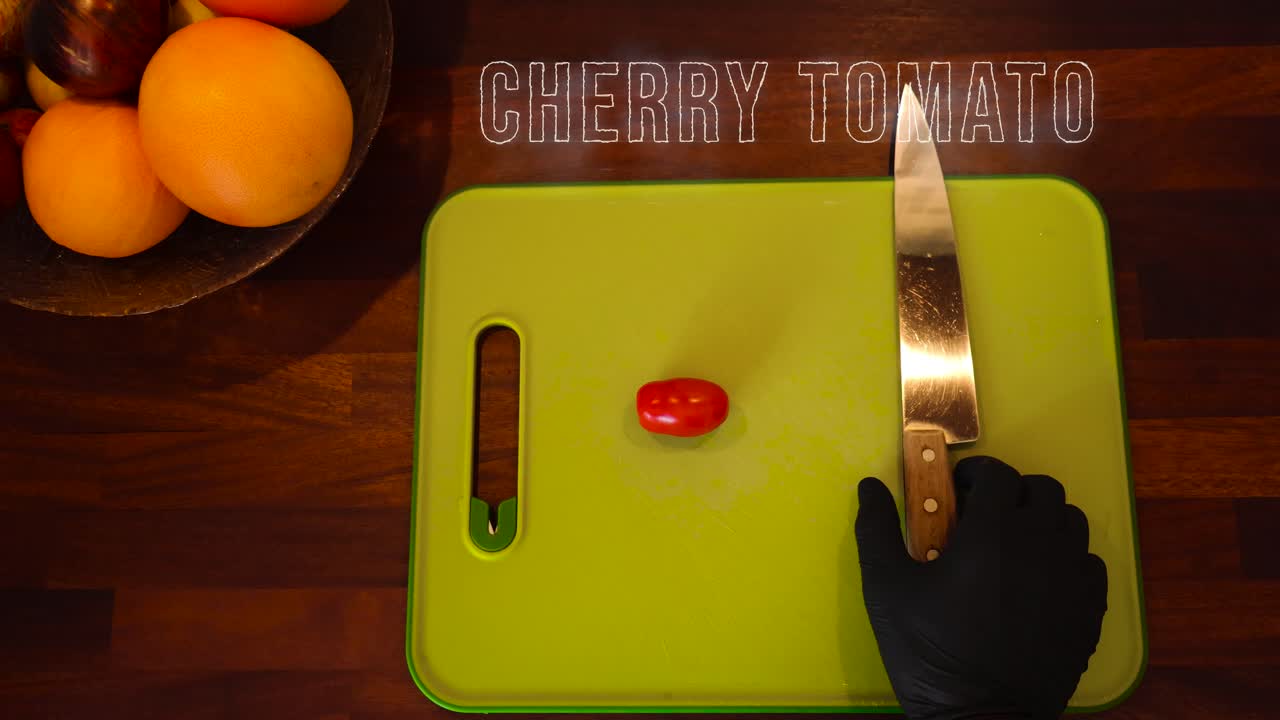 A small red cherry tomato and a knife being placed on a cutting board while a text animation on top of the kitchen counter says cherry tomato