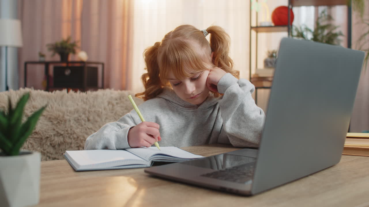 Tired young girl doing homework at table with laptop writing in notebook and showing lazy expression