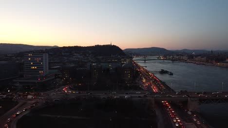 Vista-Aérea-Del-Tráfico-Por-La-Noche-Con-El-Río-Danubio-En-Budapest-Con-La-Ciudad-Y-Las-Montañas-Al-Fondo