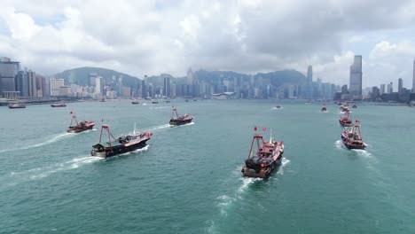 Convoy-De-Barcos-De-Pesca-Locales-Que-Causan-En-La-Bahía-Victoria-De-Hong-Kong,-Con-El-Horizonte-De-La-Ciudad-En-El-Horizonte,-Vista-Aérea