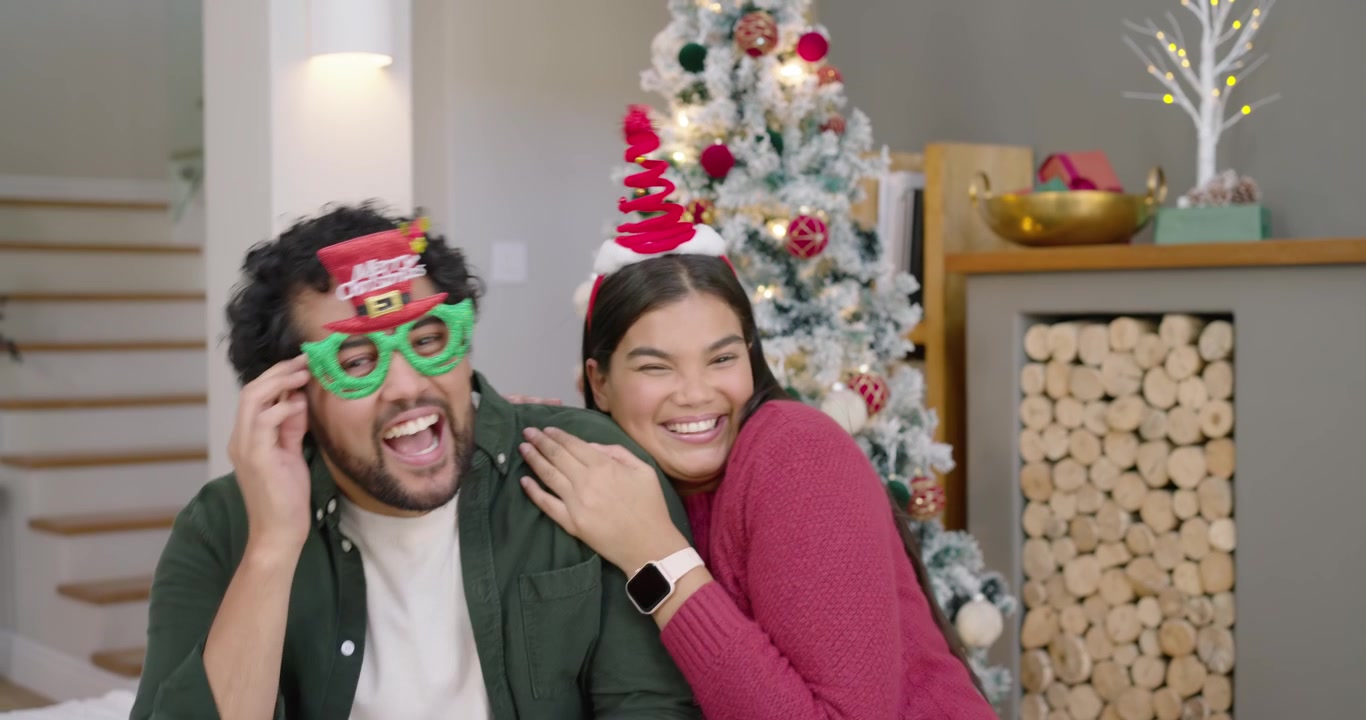 Pareja compartiendo miradas, mujer tocándolo y arreglándole la diadema, hombre poniéndose gafas verdes en casa para Navidad.