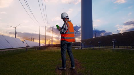 full body back view of asian male engineer with safety helmet working on a green screen smartphone and looking around while standing with solar panel and wind turbines
