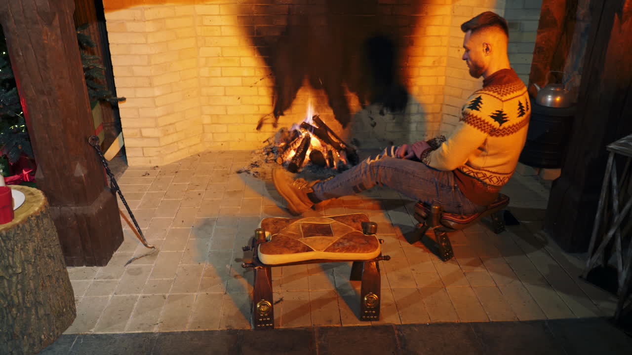 Young bearded guy sit on the chair, rubbing his hands to warm up near fire. Handsome male in sweater inside.