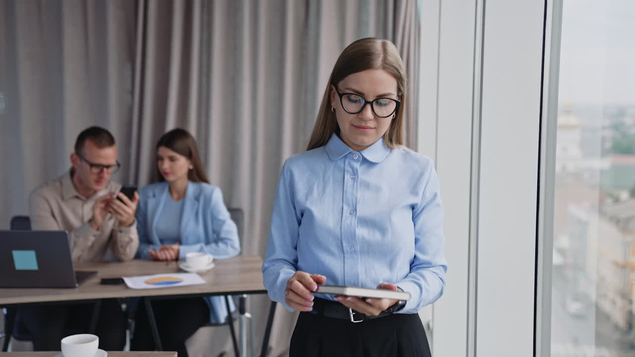 Female office employee stands at the window turning pages of her paper notebook. Colleagues sit at desk behind and talk. Blurred background.