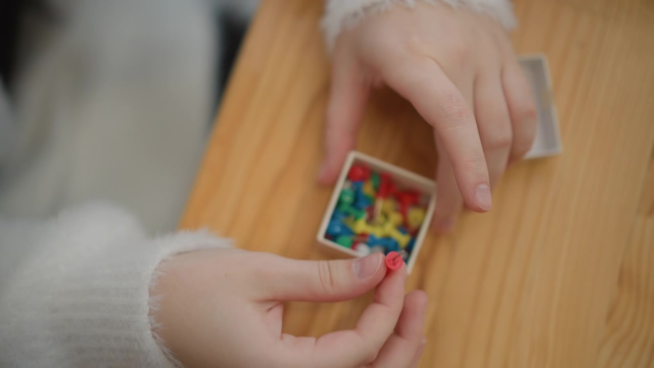 Vista ravvicinata all'altezza degli occhi di mani che inseriscono un piolo rosso in una scatola aperta di pezzi di gioco multicolori su un tavolo di legno caldo, con dita attente, presa ferma e un movimento calmo e organizzato in una luce interna accogliente.