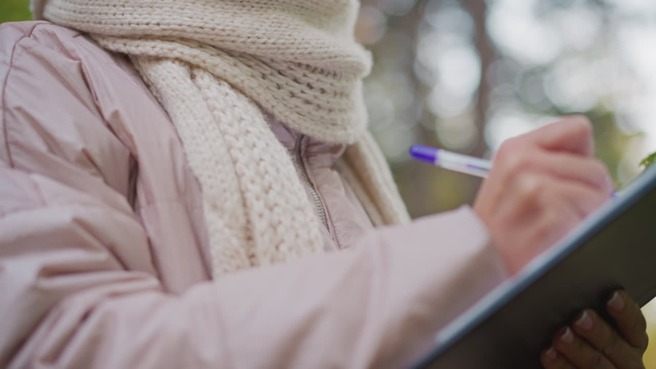 close up of mature woman in pink jacket and white scarf writing something down with gentle expression on face, surrounded by soft-focus green forest background