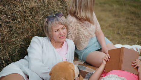 family enjoys quiet outdoor moment resting near hay bale in open countryside, thoughtful woman gazes into distance, another leans back, and child lies down reading