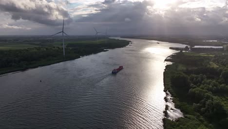 Frachtschiff-Fährt-Bei-Sonnenuntergang-Einen-Malerischen-Fluss-Hinunter,-Im-Hintergrund-Windturbinen