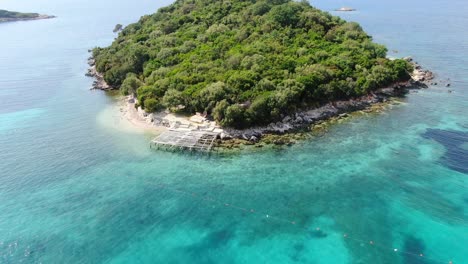 Drone-view-in-Albania-circling-in-front-of-a-green-small-island-over-crystal-clear-blue-water-ocean,-sunny-day-in-Ksamil