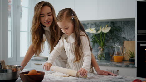 madre tierna ayudando a su hija en la cocina de cerca. adorable niño rodando la masa