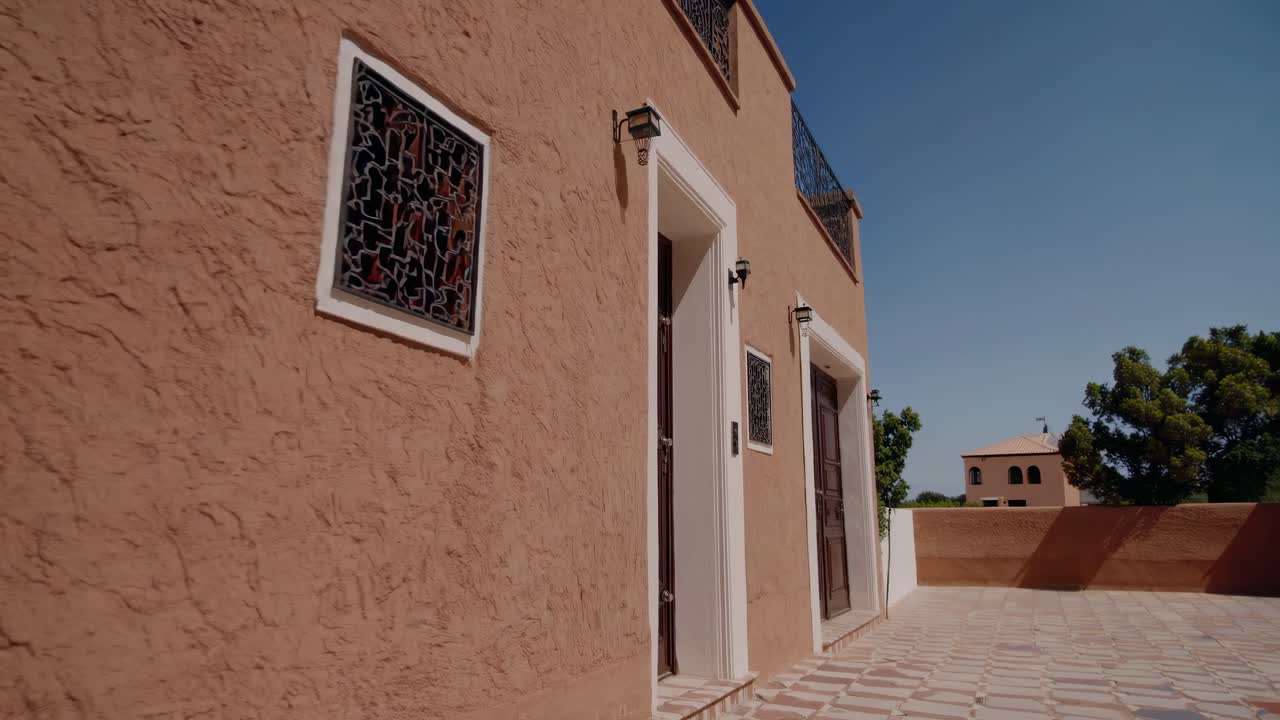 Sunlight illuminates the textured, earthen walls of a traditional Moroccan building, highlighting its ornate windows, sturdy wooden doors, and tiled terrace under a clear blue sky