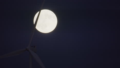 closeup silhouette - a wind turbines blades spin in front of a full moon