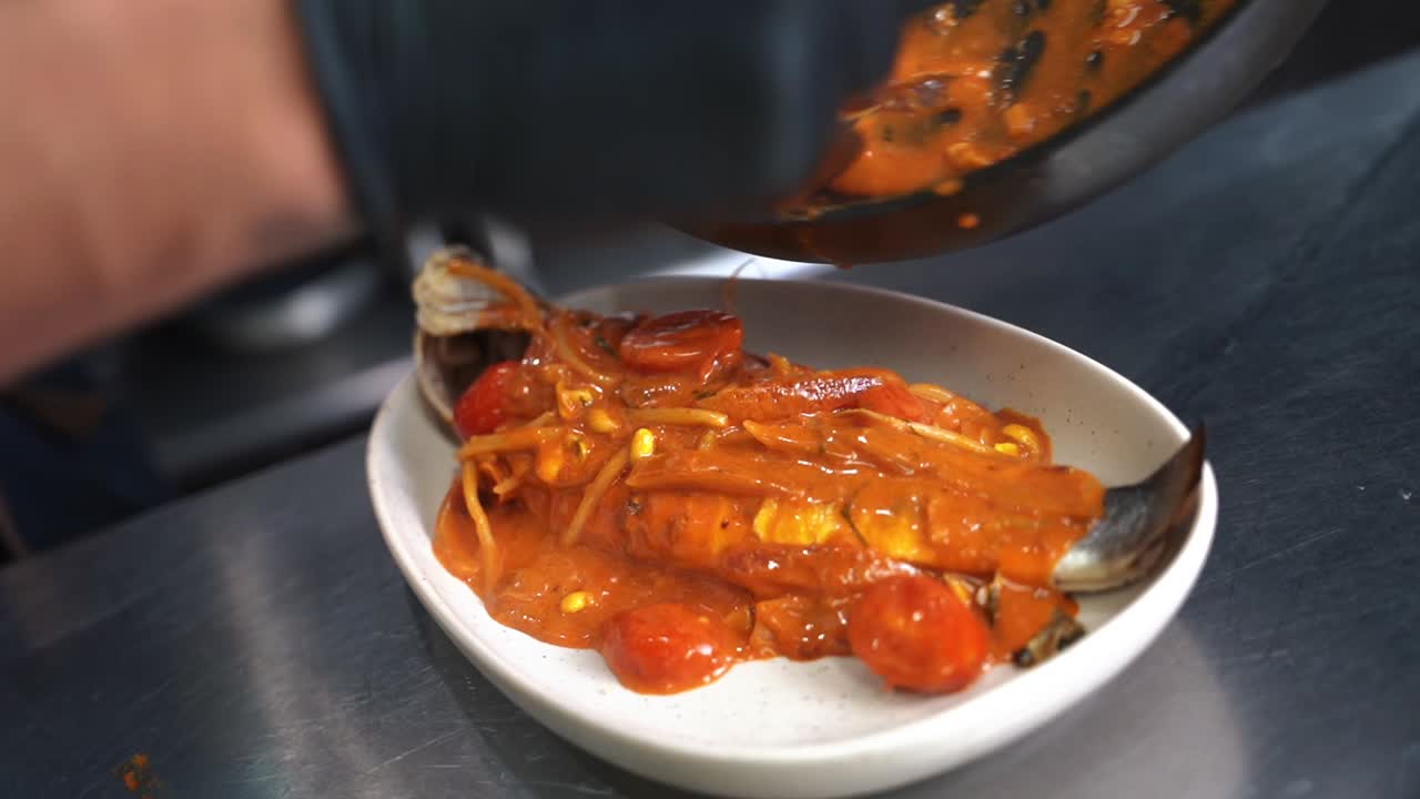 Professional chef places sauce over whole fish on a stainless steel work surface in a kitchen