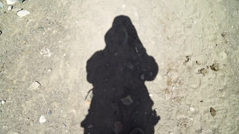 POV-shot-filming-own-shadow-as-walking-on-a-dirt-road,-a-hiker-in-nature,-with-the-feet-in-the-frame,-looking-down