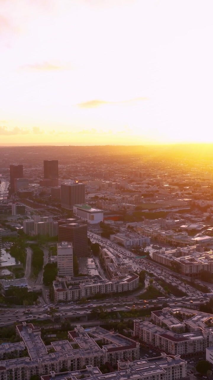 O horizonte de Los Angeles exibe arranha-céus e torres de escritórios ao pôr do sol. Congestionamento de tráfego visível nas autoestradas. Palmeiras emolduram a rede de transporte urbano no centro da cidade.