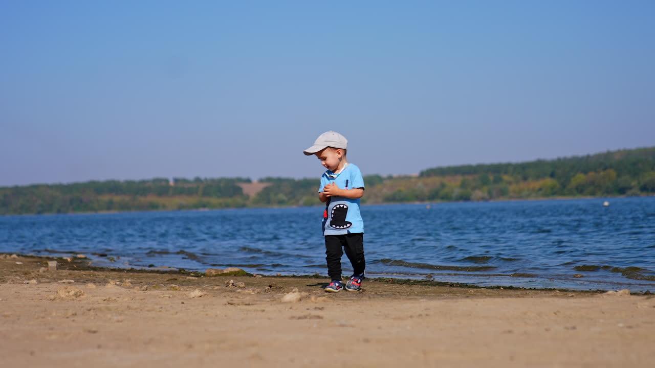 Lovely kid walks by the sandy river bank with no other people around. Toddler picks up a stone and throws it into water.