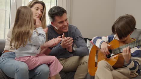 a family gathers in their living room, watching and applauding as a young boy plays an acoustic guitar