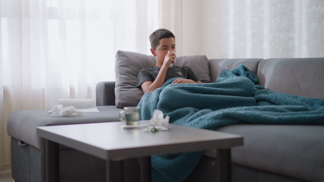 Sick little boy sitting on couch covered with blue blanket, sneezing into tissue and cleaning his nose, tissue on table with glass cup beside him. Indoor, cozy, home environment
