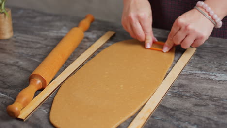 close-up of woman cutting dough using a baking tool, hands pressing orange cookie cutter, wooden rolling pin beside, dough neatly rolled, pink beaded bracelet on wrist