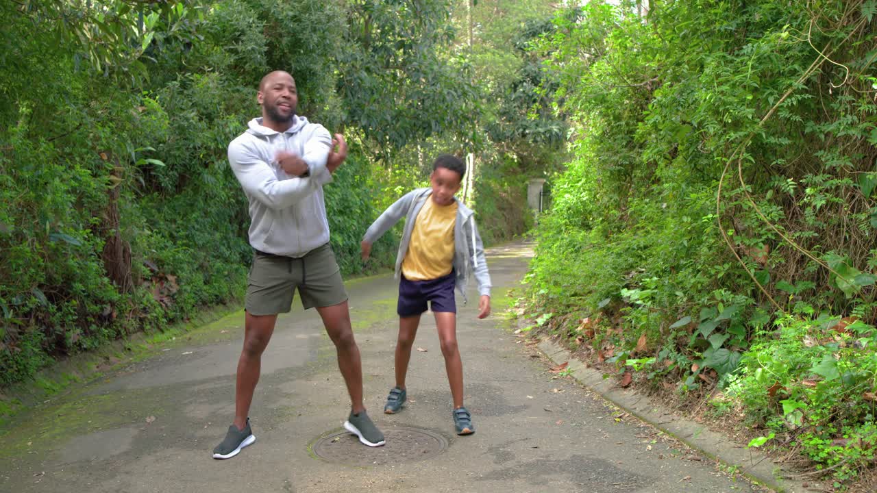 African American father and son performing warmup exercises on trail near manhole cover before run
