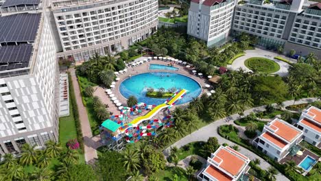 aerial footage of southeast asian resort complex shows central pool with colorful water slides and lounge areas, all amidst lush palm trees and gardens,a well-appointed, family-friendly atmosphere.