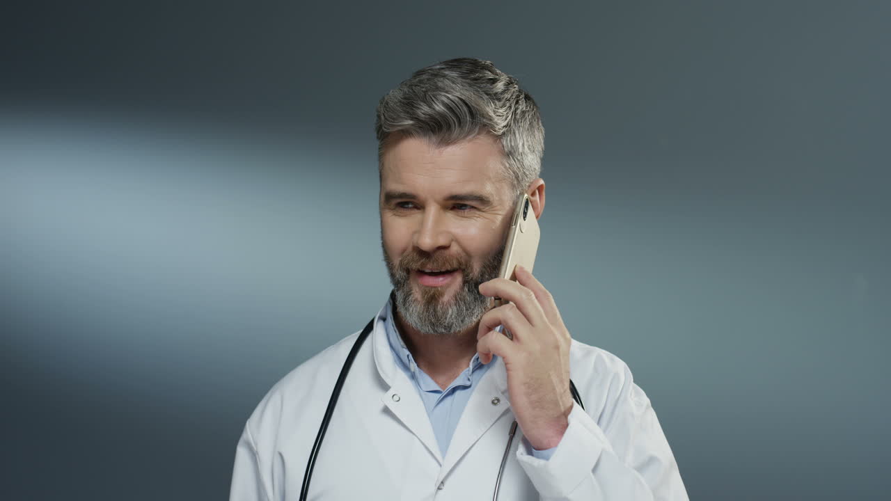 Handsome Man Doctor Talking Cheerfully On Mobile Phone And Laughing On The Gray Wall Background