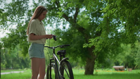 young woman with long hair approaches black bicycle parked on paved path, wears gloves with lush greenery and tall trees in background