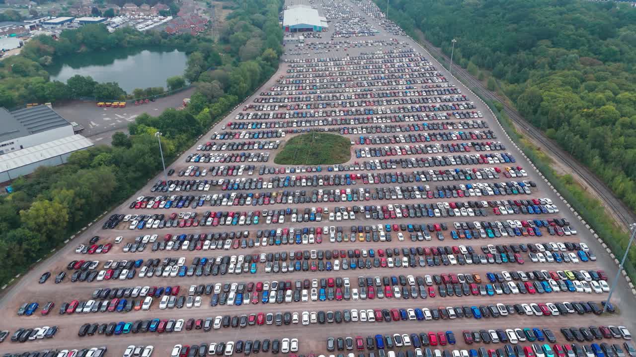A large car park with neatly arranged cars in various colors, surrounded by greenery and a water body, with an urban area in the background. The scene captures the organized and structured layout.