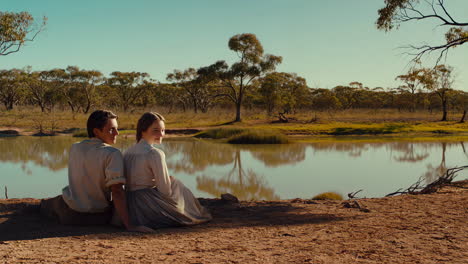 a couple sits by a tranquil lake in a vast natural landscape