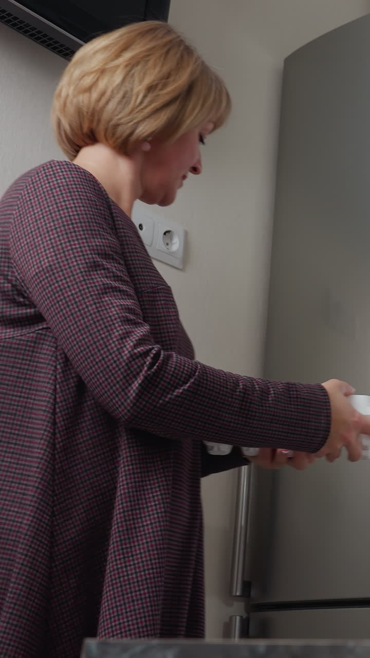 mujer abre la nevera para recuperar una caja de huevos mientras está de pie cerca de un enchufe de pared y la televisión, luego cierra la nevera, se aleja, y coloca los huevos en la encimera en una cocina moderna