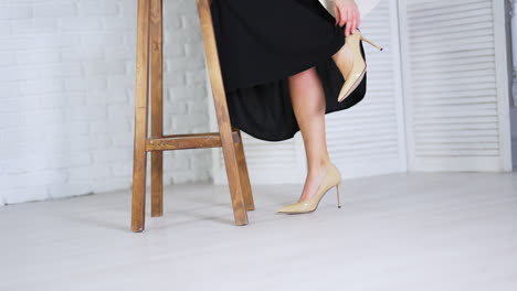 caucasian woman in beige stiletto shoes comes up to a wooden tabouret. model lifts her leg to feel tired foot from walking on high heels.