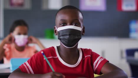 portrait of african american schoolboy wearing face mask, sitting in classroom looking at camera