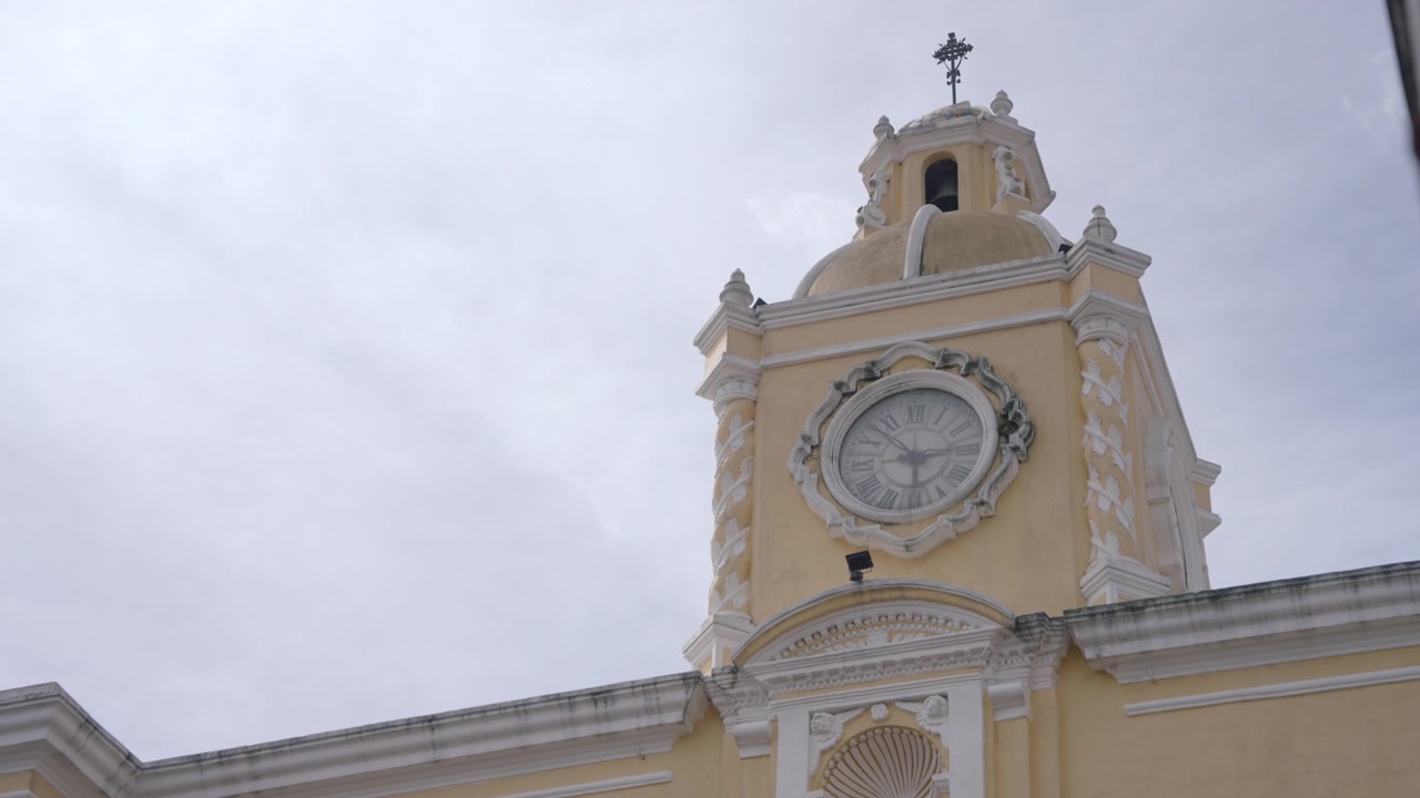 Low-angle view of a historic yellow building, Arco de Santa Cantalina, with a prominent clock tower against an overcast sky in Antigua, Guatemala