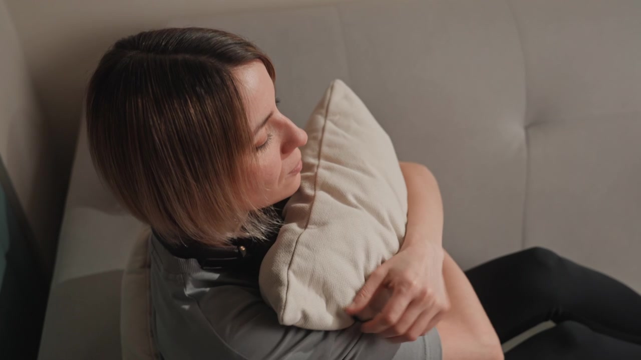 Giovane donna sul divano che abbraccia un cuscino, amante della musica con le cuffie al collo. Un umore tranquillo e riflessivo. Morbido divano grigio, angolazioni ravvicinate, un gesto tenero, un'espressione malinconica e un bagliore accogliente della lampada.