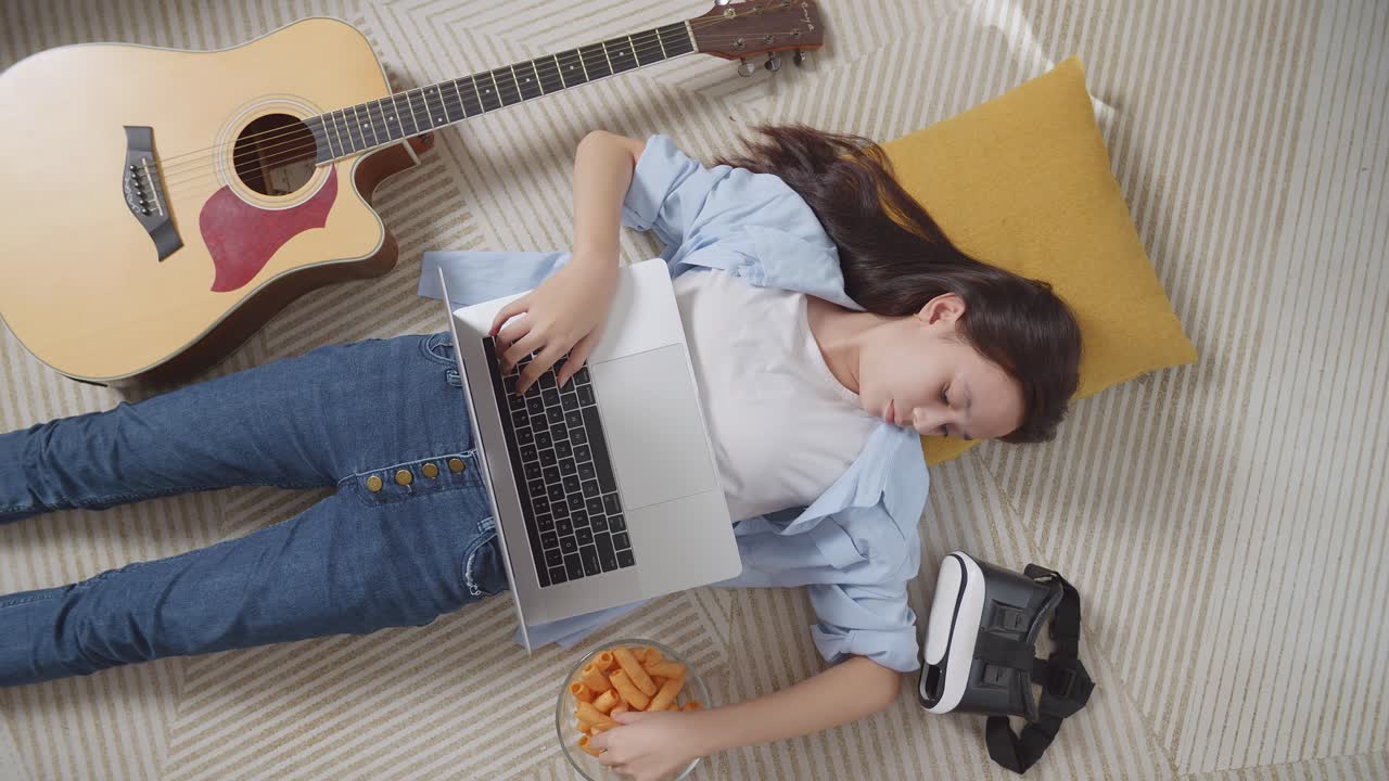 vista superior de una adolescente asiática comiendo bocadillos y escribiendo en una computadora portátil mientras yacía en la alfombra en el suelo de su casa