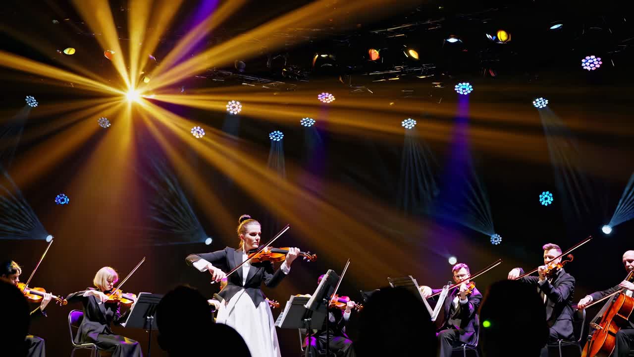 Professional musicians playing violins in an orchestra during a live concert, surrounded by vibrant stage lights, create a captivating and magical atmosphere for the audience