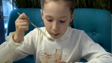 close up. a little girl with freckles on her face, in a white dress, with pigtails, sits and eats in a cafe with a spoon dessert made of a glass vase. portrait.