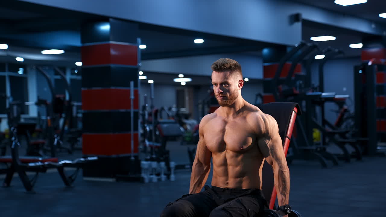 Bell'atleta di forza, bodybuilder, esegue esercizi con i manubri. Corpo muscoloso e in forma su sfondo scuro di palestra.