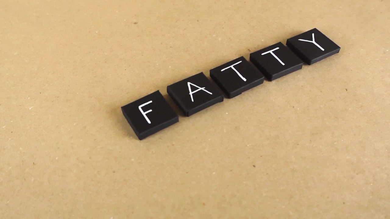 Filmagem em panorâmica lenta sobre a palavra "fatty" soletrada com peças de letras quadradas pretas em um fundo liso marrom claro, representando conceitos de dieta não saudável e obesidade