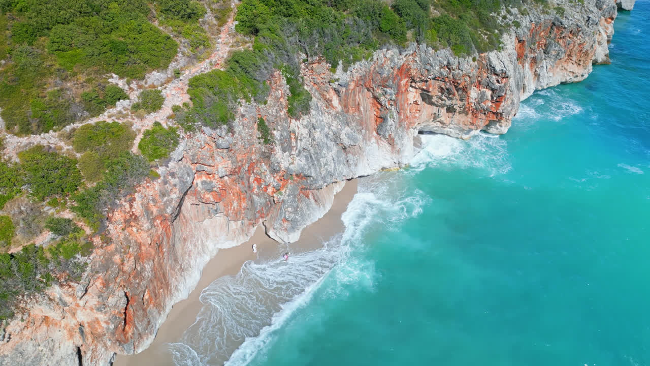 Ripresa aerea con zoom di un drone sulle onde che si infrangono sulla spiaggia di Gjipe, Albania, lungo una ripida scogliera in una giornata di sole.