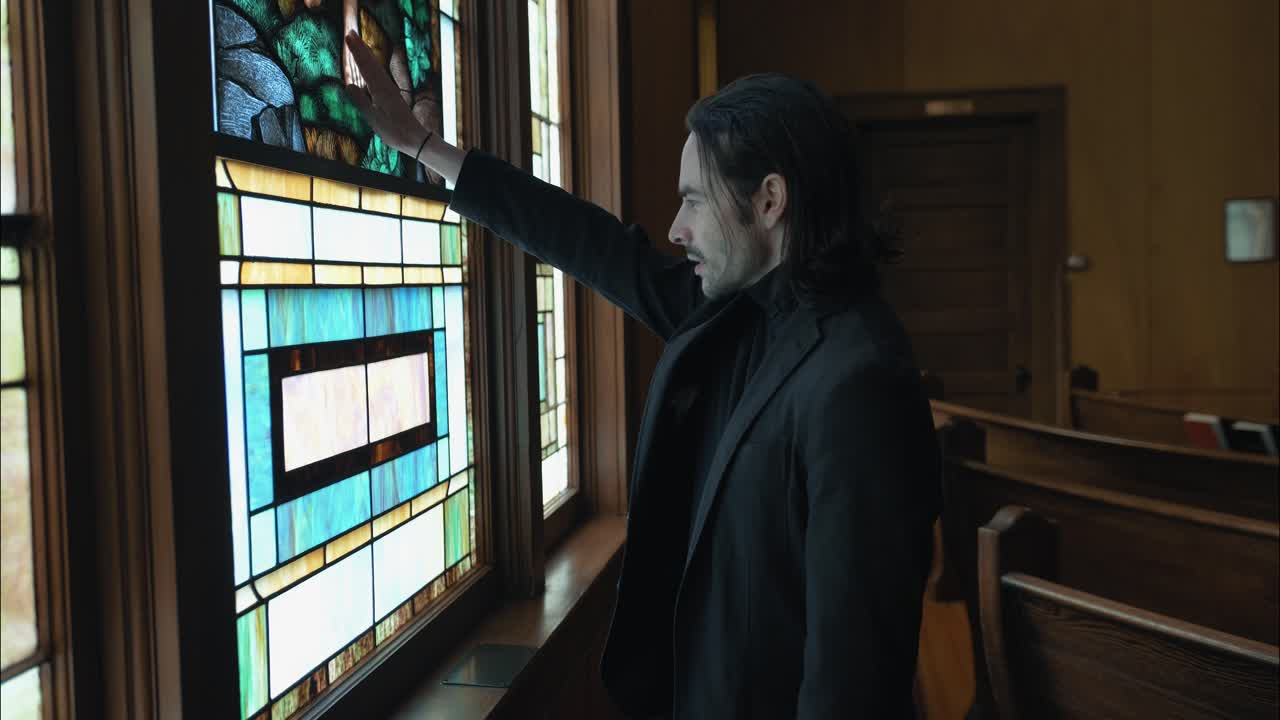 A lone figure stands in the pews of a church, facing a stained-glass window, symbolizing prayer, contemplation, and the search for meaning