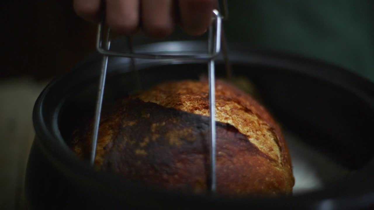 Hot and freshly baked bread loaf lifted out of baking pot with metal tool, filmed as closeup slow motion shot