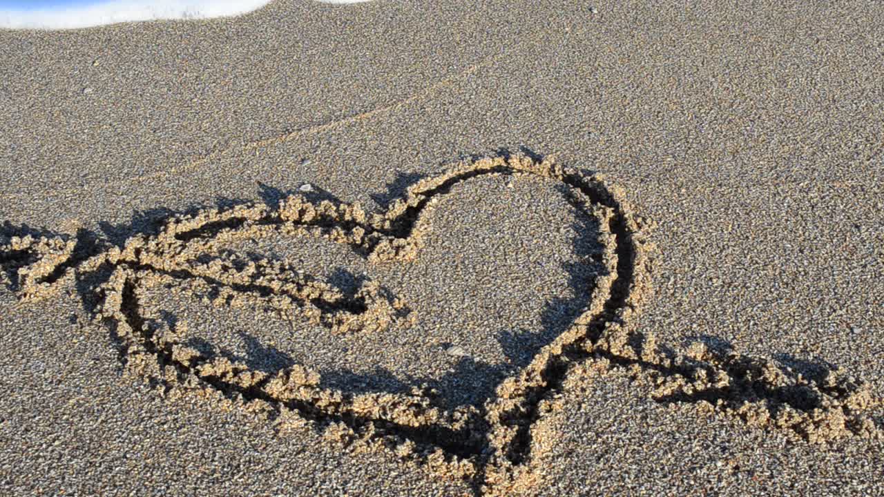 Un cuore disegnato sulla sabbia. Il cuore viene cancellato da un'onda marina. Riprese sulla spiaggia.